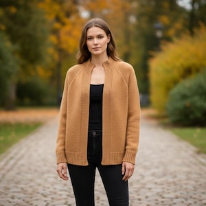 May include: A woman wearing a tan cardigan sweater over a black top and black pants. The open-front cardigan has a ribbed texture and long sleeves. The woman is standing on a stone path in a park with autumn foliage.