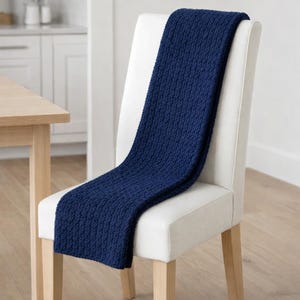 May include: A navy blue knitted throw blanket draped over the back of a white upholstered chair. The blanket has a textured pattern and is made of a soft material. The chair has wooden legs and is in a well-lit room.