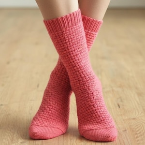 May include: A pair of coral-coloured knitted socks, with a textured pattern, are crossed at the ankles. The socks are made of a thick yarn and have ribbed cuffs. The background is a light-coloured wooden floor.