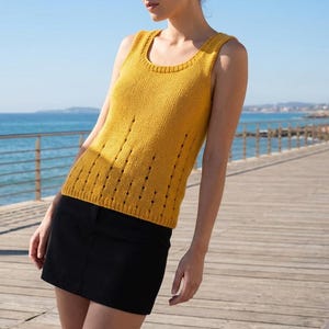 May include: Mustard yellow knitted vest top with a scoop neckline and vertical cut-out detailing. The top is worn with a black mini skirt. The background shows a wooden boardwalk, the sea, and a blue sky.