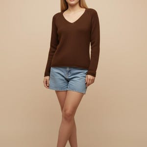 May include: A woman wearing a long-sleeved, dark brown V-neck sweater and light blue denim shorts. The sweater has a simple design, and the shorts are a classic style. The background is a solid, neutral color.