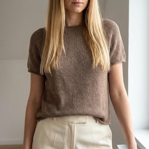 May include: A short-sleeved, brown knit top with a crew neck. The top is paired with cream-colored pants. The model has long blonde hair. The clothing is likely handmade and suitable for casual wear.