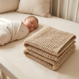 Könnte beinhalten: Ein Stapel von drei gefalteten, beigefarbenen Strickdecken. Die Decken bestehen aus dickem, strukturiertem Garn. Im Hintergrund schläft ein Baby, das in eine weiße Decke gewickelt ist, in einem Kinderbett.