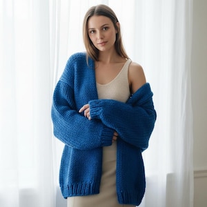 Puede incluir: Una mujer con un cárdigan de punto azul cobalto grueso y extragrande. El cárdigan tiene un corte holgado con mangas largas. Debajo, se ve una camiseta sin mangas y un vestido beige. El fondo presenta una cortina blanca.