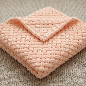 May include: A folded, peach-colored blanket with a woven texture. The blanket is made of a soft, plush material and is folded into a square shape. The top corner is folded over, creating a layered effect. The blanket is on a neutral-colored surface.