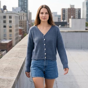 May include: A woman wearing a blue cardigan with white buttons and denim shorts. The cardigan has a V-neck and long sleeves. The background features a cityscape with tall buildings.