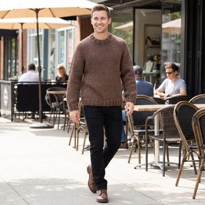 May include: A man wearing a brown knit sweater and black pants walks down a sidewalk. He is wearing brown leather shoes. The background includes outdoor seating, umbrellas, and storefronts.