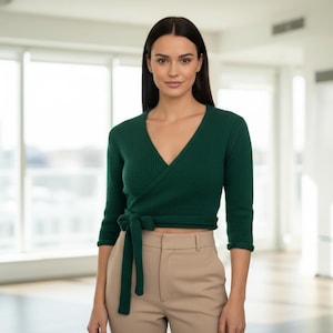 May include: A woman wearing a dark green wrap sweater with a V-neck and three-quarter sleeves. The sweater is tied at the waist. She is wearing beige pants. The background is a bright, modern interior.