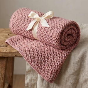 May include: A rolled-up, dusty rose-colored knitted blanket with a woven pattern, secured with a cream-colored satin ribbon tied in a bow. The blanket is partially unfolded, resting on a wooden surface and a linen cloth.