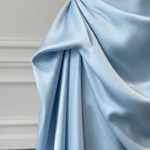 May include: A close-up of a light blue satin dress. The fabric is draped and gathered, creating soft folds and texture. The dress appears to be a formal gown, with a smooth, elegant finish.