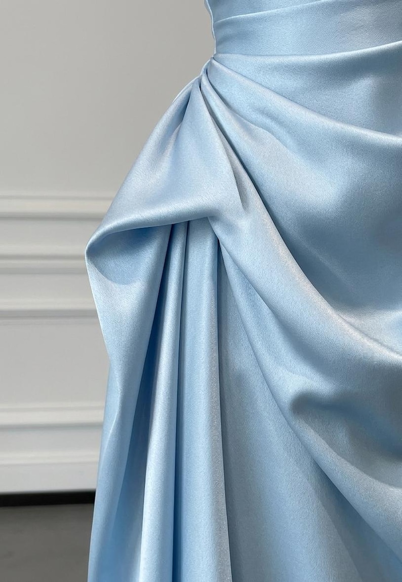 May include: A close-up of a light blue satin dress. The fabric is draped and gathered, creating soft folds and texture. The dress appears to be a formal gown, with a smooth, elegant finish.