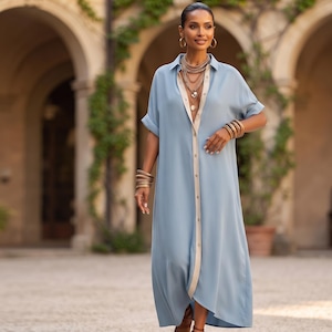 May include: A light blue button-down shirt dress featuring a contrasting beige placket, a classic collar, and short sleeves with rolled cuffs. The garment is styled with layered gold-toned necklaces and stacked gold-toned bracelets on both wrists.