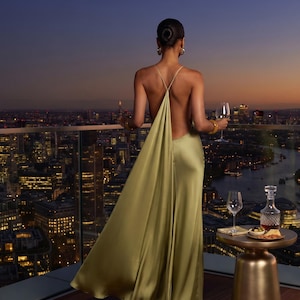 May include: A floor-length, olive green satin gown featuring a backless design with thin, delicate straps that cross at the upper back. The fabric has a smooth, lustrous finish that drapes down to the floor, creating a fluid and elegant silhouette.