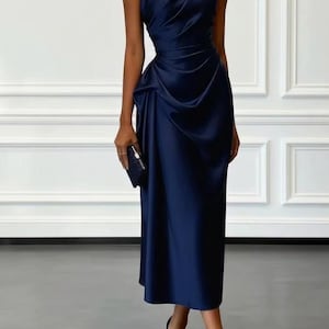 May include: A navy blue, one-shoulder midi dress with draped fabric detailing. The dress features a fitted waist and a flowing skirt. The model is wearing matching navy blue heels and carrying a small clutch. The background is a white wall with decorative molding.