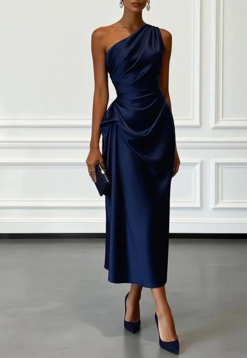 May include: A navy blue, one-shoulder midi dress with draped fabric detailing. The dress features a fitted waist and a flowing skirt. The model is wearing matching navy blue heels and carrying a small clutch. The background is a white wall with decorative molding.