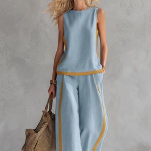 Può includere: Un top senza maniche azzurro e pantaloni larghi abbinati con profili giallo senape lungo le cuciture laterali e l'orlo. L'outfit è completato da una borsa tote a righe marroni e nere e da sandali marroni per un look coordinato.