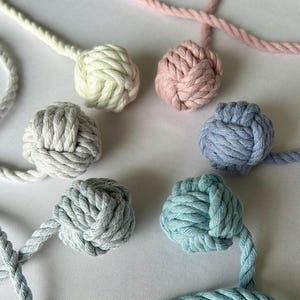 May include: A selection of decorative rope knots in pastel shades of white, pink, grey, and blue. Each knot is connected to a length of matching rope. The knots are tightly woven and appear handmade. The image is a close-up, highlighting the texture and colours.