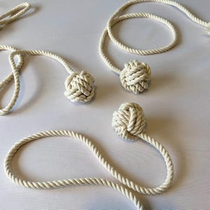 May include: Off-white rope with decorative knotted balls. The rope is arranged in loops and connected to the knotted balls. The balls are tightly woven, and the rope has a twisted texture. The background is a plain white surface.