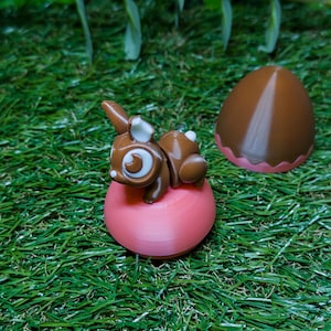 Könnte beinhalten: Eine braune Hasenfigur mit weißen Akzenten liegt auf einer rosa Kugel, neben einem braunen, eiförmigen Objekt. Die Objekte befinden sich auf grünem Kunstgras, was ein Frühlings- oder Osterthema suggeriert.
