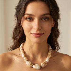 Peut inclure: Un collier composé de perles lisses et polies dans des tons crème, beige et marron avec un motif marbré. Les perles sont de forme ovale et arrondie, séparées par de petites entretoises en métal argenté, formant un design circulaire continu.