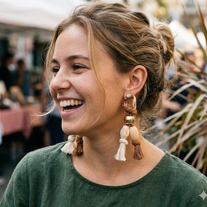Peut inclure: Une femme porte de grandes boucles d'oreilles faites à la main. Les boucles d'oreilles présentent une combinaison de matériaux, notamment des perles de bois, des accents métalliques dorés et de la corde nouée. Des glands crème et marron complètent le design. La femme porte un haut vert.