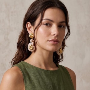 Peut inclure: Boucles d'oreilles coquillage dorées et crème. Les boucles d'oreilles présentent une boule dorée en haut, une coquille en spirale crème et un motif coquillage doré en bas. La personne porte un haut vert.