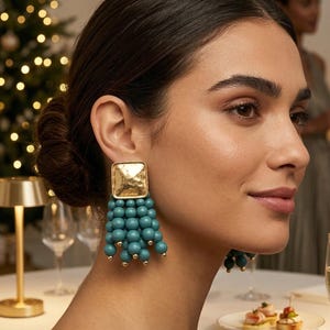 Peut inclure: Boucles d'oreilles carrées dorées avec une surface texturée, ornées d'une cascade de perles bleu sarcelle. Les boucles d'oreilles ont de petits accents dorés au bas des grappes de perles. La femme porte les boucles d'oreilles.