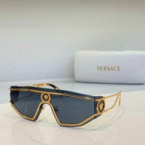 Peut inclure: Lunettes de soleil gris foncé avec monture dorée et logo Versace. Les lunettes ont un design angulaire unique. Un étui blanc avec le logo Versace est également visible.