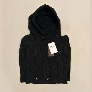 Può includere: Felpa con cappuccio nera con motivo ripetuto. La felpa ha una coulisse e un'etichetta con la scritta "Dior". Il tessuto sembra essere un materiale morbido.
