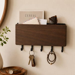 Peut inclure: Un organisateur mural en bois brun foncé avec un dessus en métal noir et quatre crochets noirs. L'organisateur contient un magazine, une enveloppe et des lunettes de soleil. Des clés et un bracelet sont suspendus aux crochets. Le magazine indique "CEREAL".