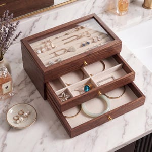 Puede incluir: Una caja de joyería de madera oscura con tapa de cristal y varios cajones. La caja contiene collares, pendientes y pulseras. Los cajones están forrados con tela beige. Los cajones tienen pomos dorados.