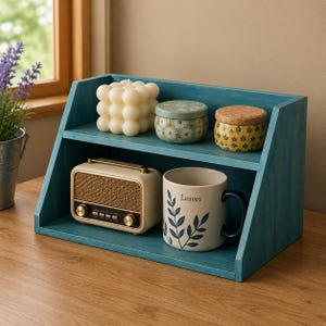 May include: A teal wooden two-tier shelf holding a white bubble candle, a floral patterned tin, a yellow patterned tin with a cork lid, a beige retro-style radio with gold knobs, and a white ceramic mug with blue leaves and the text Leaves.