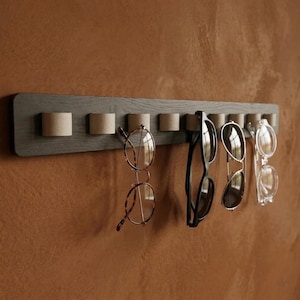 Puede incluir: Un organizador de gafas de madera gris con varios pares de gafas y gafas de sol colgadas. El organizador tiene una serie de pequeños soportes cilíndricos. Las gafas incluyen monturas redondas, cuadradas y de ojo de gato en varios colores.