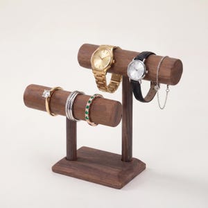 May include: A wooden jewellery display stand with two levels. The top level holds a gold watch, a silver watch with a black strap, and a silver chain bracelet. The bottom level displays a gold ring, silver rings, and a ring with green stones.