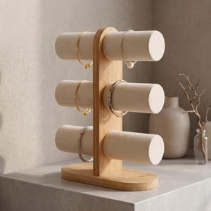 Op de afbeelding: Een sieradenstandaard met drie niveaus, gemaakt van lichtgekleurd bamboe. De standaard heeft drie cilindrische, crèmekleurige, gewatteerde staven voor het presenteren van armbanden. Er worden verschillende armbanden in goud- en zilverkleuren getoond.