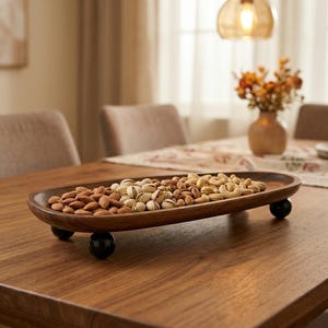 以下が含まれることがあります： アーモンド、ピスタチオ、カシューナッツが盛られた木製のサービングトレイ。 楕円形のトレイは濃い茶色で、3つの小さな黒い球形の足の上に置かれています。 トレイは木製のテーブルの上に置かれています。