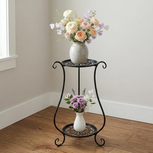 May include: A black metal plant stand with two circular shelves, each holding a vase of flowers. The top shelf has a gray vase with peach and lavender flowers. The bottom shelf has a white vase with purple and white flowers.