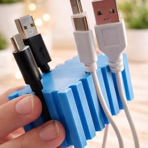 May include: A blue 3D-printed cable organizer holds several USB cables. The cables are black and white, with various connector types. The organizer is held in a hand, and the background is blurred.