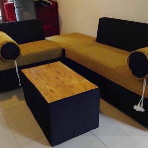 Könnte beinhalten: Ein modernes Ecksofa und ein passender Couchtisch. Das Sofa hat einen schwarzen Stoff mit senffarbenen Kissen und zylindrischen Nackenrollen. Der Couchtisch hat eine Holzplatte und schwarze Seiten. Die Möbel stehen in einem hellen Raum.