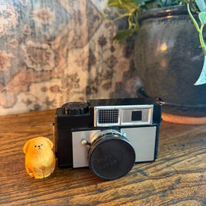May include: A vintage black and silver camera with a lens cap and a small wooden dog figurine on a wooden surface. The camera has a rectangular viewfinder and a round lens. The dog is light brown and carved.
