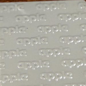 May include: A square, off-white tile or surface featuring a repeating, embossed pattern of the word apple in lowercase letters. The text is raised and integrated into the material, creating a subtle, monochromatic texture across the entire surface.