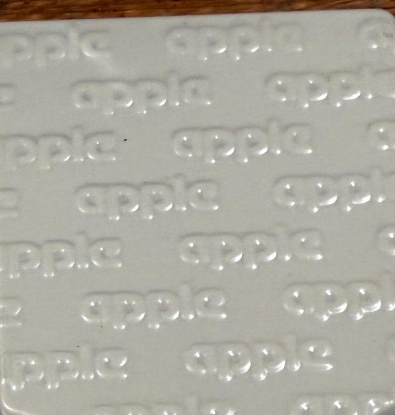 May include: A square, off-white tile or surface featuring a repeating, embossed pattern of the word apple in lowercase letters. The text is raised and integrated into the material, creating a subtle, monochromatic texture across the entire surface.