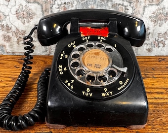 May include: A vintage black rotary telephone with a coiled cord and a circular dial featuring white letters and numbers. The center of the dial contains a circular paper insert with the text AREA 703 and 243-8867, and a red label above reads DIAL 911.