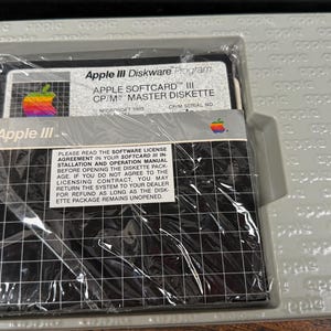 May include: A vintage Apple III Softcard CP/M master diskette in its original grey plastic tray. The black diskette features a white grid pattern, a rainbow Apple logo, and text reading Apple III Diskware Program and Apple Softcard III CP/M.