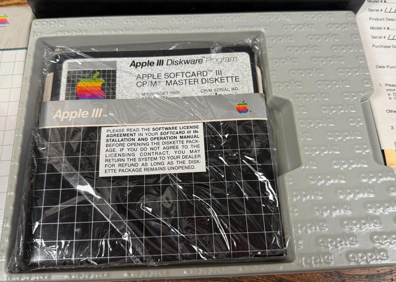 May include: A vintage Apple III Softcard CP/M master diskette in its original grey plastic tray. The black diskette features a white grid pattern, a rainbow Apple logo, and text reading Apple III Diskware Program and Apple Softcard III CP/M.