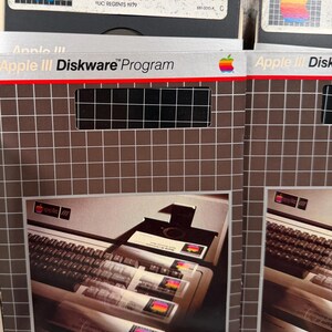Puede incluir: Dos fundas de software Apple III Diskware Program con un patr&oacute;n de cuadr&iacute;cula y un logotipo de manzana arco&iacute;ris. Las fundas muestran una fotograf&iacute;a de un teclado y unidad de disco de computadora Apple III. El texto incluye Apple III Diskware.