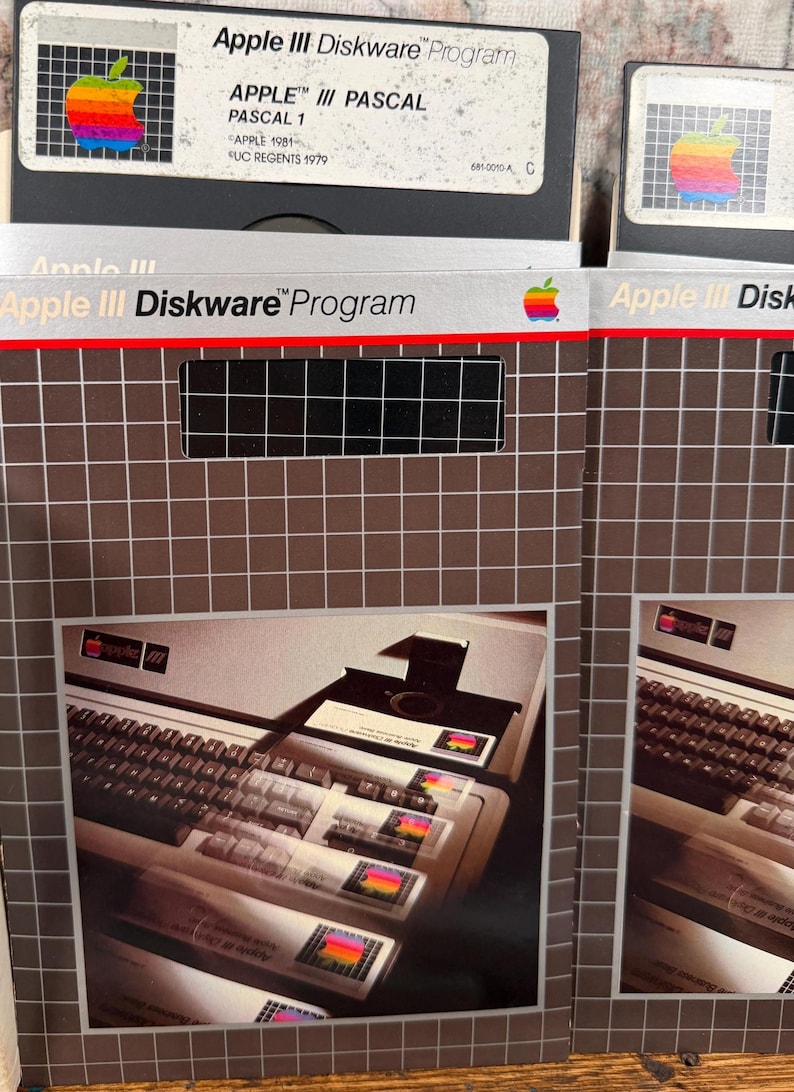 Puede incluir: Dos fundas de software Apple III Diskware Program con un patr&oacute;n de cuadr&iacute;cula y un logotipo de manzana arco&iacute;ris. Las fundas muestran una fotograf&iacute;a de un teclado y unidad de disco de computadora Apple III. El texto incluye Apple III Diskware.
