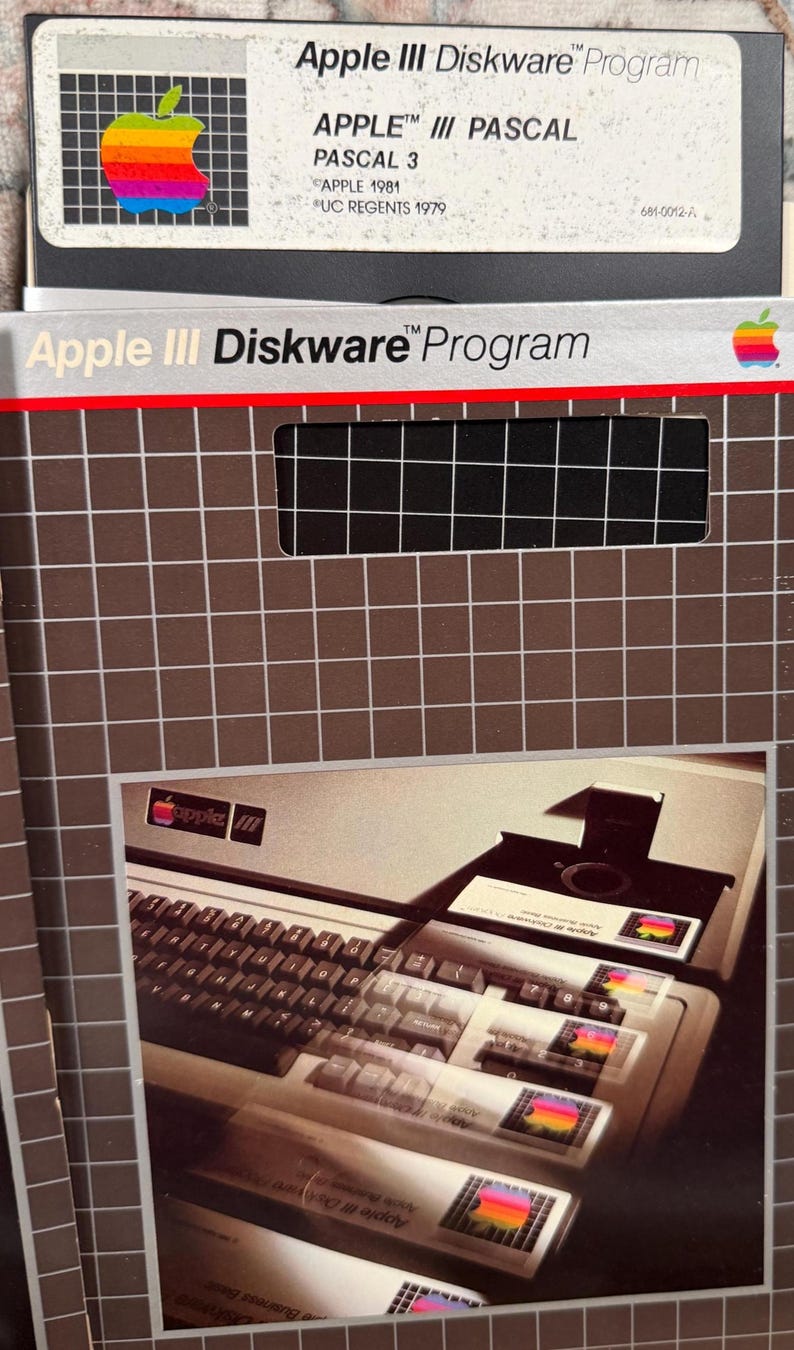 Puede incluir: Un disquete vintage Apple III Diskware Program y su funda de cart&oacute;n. La funda presenta un patr&oacute;n de cuadr&iacute;cula marr&oacute;n, el texto Apple III Diskware Program y una foto de un teclado de computadora Apple III.