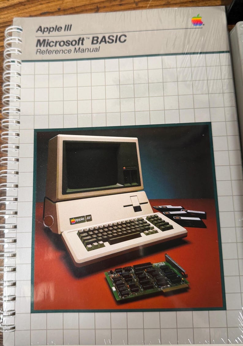 May include: A spiral-bound Apple III Microsoft BASIC Reference Manual with a white cover featuring a grid pattern. The cover displays an illustration of an Apple III computer, several floppy disks, and a circuit board on a dark red surface.