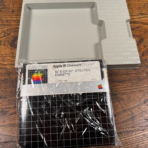 May include: A vintage Apple III Diskware Program SOS-CP/M Utilities diskette in its original plastic wrap, featuring a black grid pattern and the classic rainbow Apple logo. It is accompanied by a light grey plastic storage tray with Apple branding.